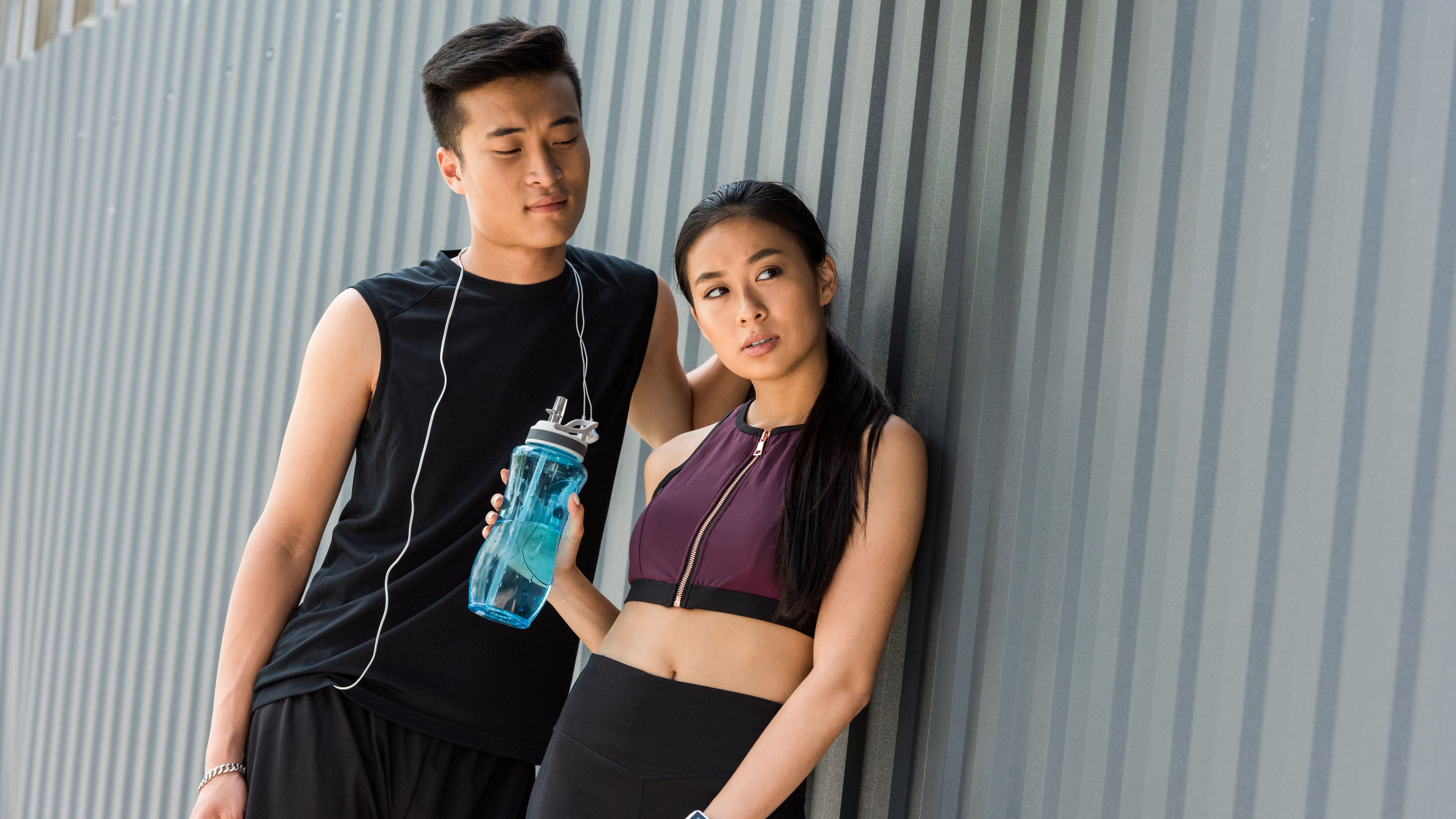 Smiling young Asian sportsman with earphones over neck standing near sportswoman with sport bottle